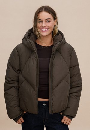 Junge Frau lächelt und trägt eine wattierte dunkelbraune Kapuzenjacke über einem dunklen Oberteil und blauen Jeans vor einem schlichten beigen Hintergrund.