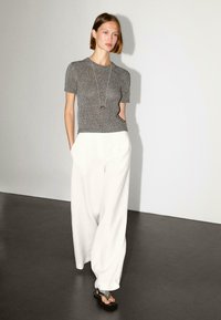 Gray knit top with short sleeves, paired with loose white trousers. The model wears a long necklace and black sandals. Simple background.