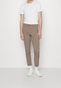 Pantaloni slim-fit marroni realizzati in tessuto liscio, con tasca anteriore e chiusura con bottone, abbinati a una t-shirt bianca semplice e sneakers bianche.