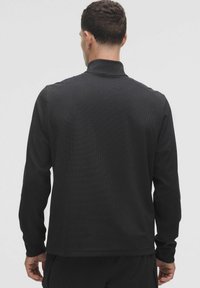 Juodas ilgomis rankovėmis polo marškinėliai su tekstūruotu raštu, turintys stovinčią apykaklę ir rankogalius. Pozicijoje, rodant galinį vaizdą, demonstruojamas pritaikytas kirpimas.