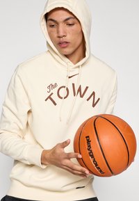 Krémová mikina s kapucí s nápisem "The TOWN" v hnědé barvě, kapuce na šňůrky, oblečená osoba držící oranžový basketbalový míč s černým nápisem.