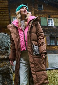 Kobieta w błękitnej czapce, włożona w brązowy i różowy płaszcz, stoi na zewnątrz obok kamiennego muru, w tle drewniany budynek, w lekkim deszczu.