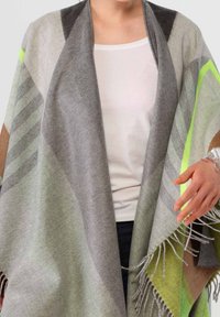 Personne portant un châle à motifs géométriques multicolores avec des franges par-dessus un haut blanc, la main ajustant le tissu.