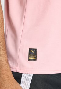 T-shirt de football rose évacuant l'humidité avec un tissu texturé et une étiquette noire arborant le logo Puma, indiquant "FOOTBALL LICENCIÉ AUTHENTIQUE".