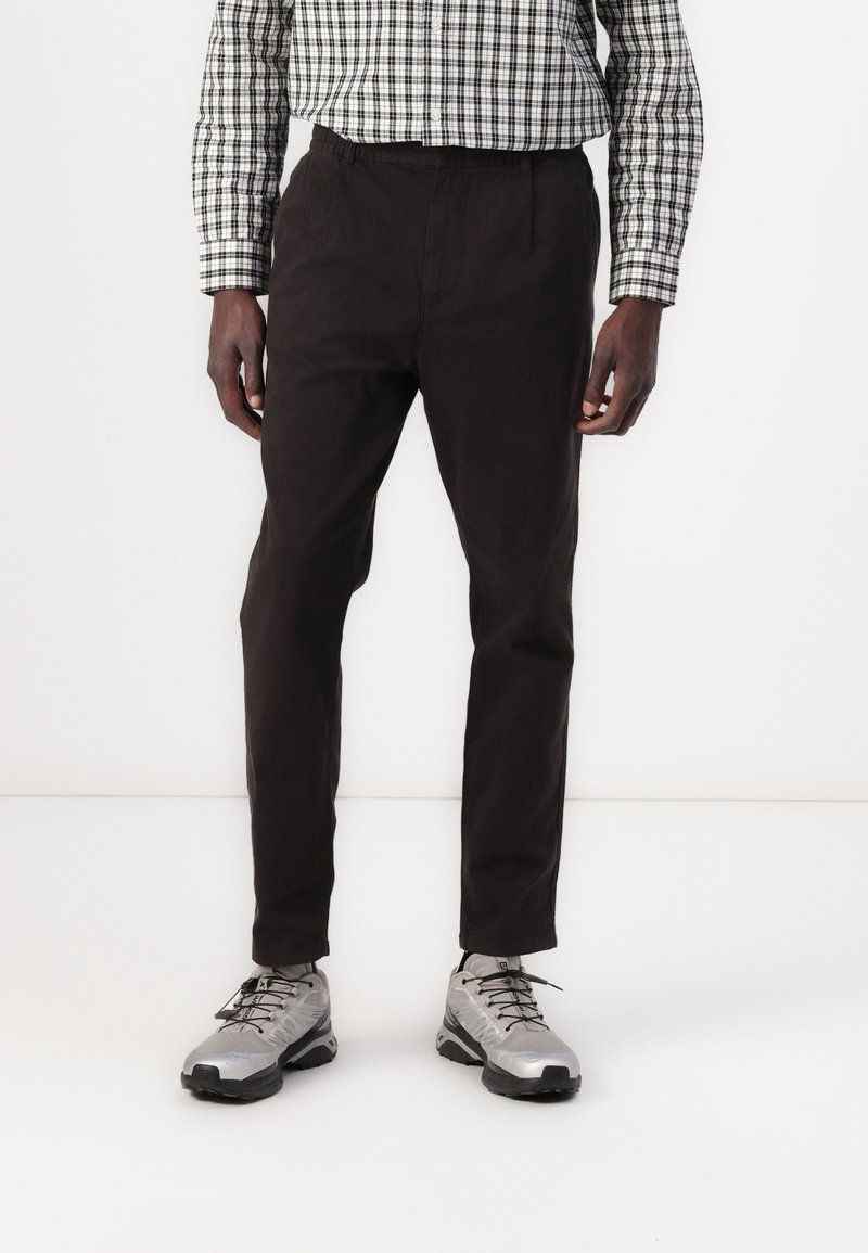Homme portant un pantalon noir sur mesure, des chaussures de sport argentées et une chemise à manches longues à carreaux noir et blanc, debout sur un fond blanc.