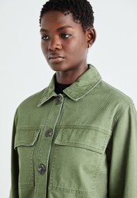 Grønn denimjakke med krage, to brystlommer og store knapper. Stoffet har en teksturert finish.
