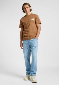 Brun bomullströja med vit "LEE" logotyp på vänster bröst, i kombination med ljusblå denimjeans och beige träningsskor.