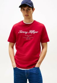 Rød bomulds T-shirt med hvid "Tommy Hilfiger" logo tekst, rund halsudskæring, korte ærmer, båret med blå jeans. Marineblå kasket på modellen.
