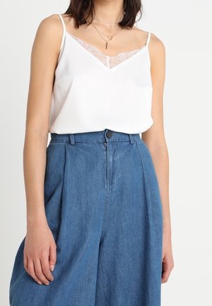 Vrouw draagt een wit kanten hemdje, ingestopt in een blauw, wijduitlopend, hooggetailleerd spijkerbroek, met gelaagde gouden kettingen.