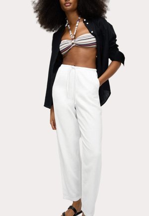 Kvinde iført sort åben skjorte, mønstret halter bandeau-top, hvide bukser med snøre og sorte sandaler, poserer med den ene hånd i lommen.