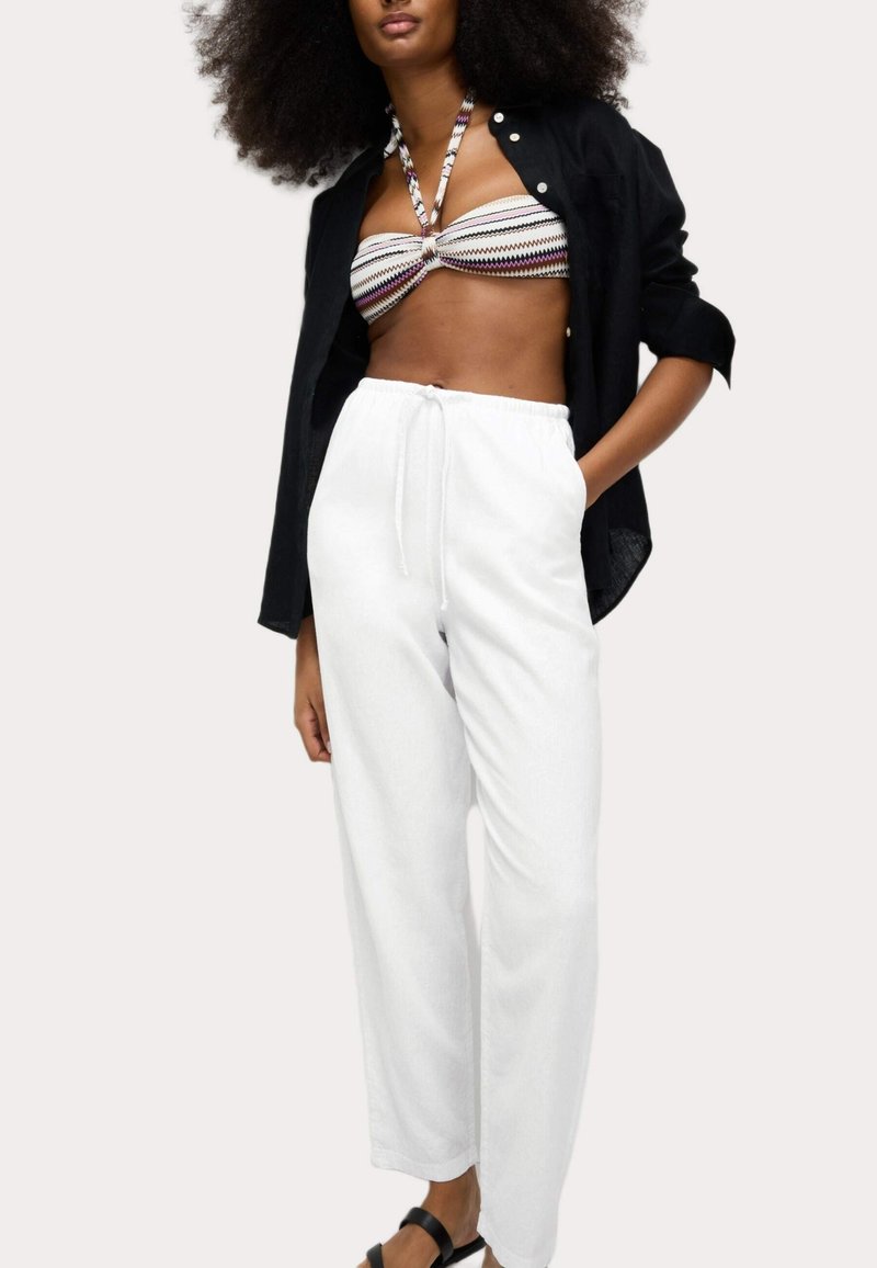 Femme portant une chemise noire ouverte, un haut bandeau à motif à col halter, un pantalon blanc à cordon de serrage et des sandales noires, posant avec une main dans la poche.