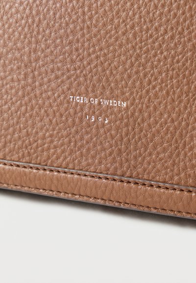 Καφέ υφή δερμάτινης επιφάνειας με ανάγλυφη τη φράση «TIGER OF SWEDEN 1903» σε λευκό, με ορατή ραφή κατά μήκος της άκρης.