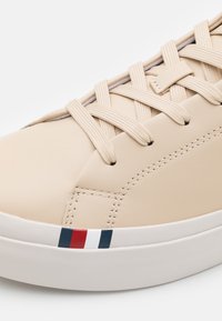 Beige lädersneaker med platta beige skosnören, diskret sömnad och en vit sula med röd, vit och blå randdekorationer på sidan.