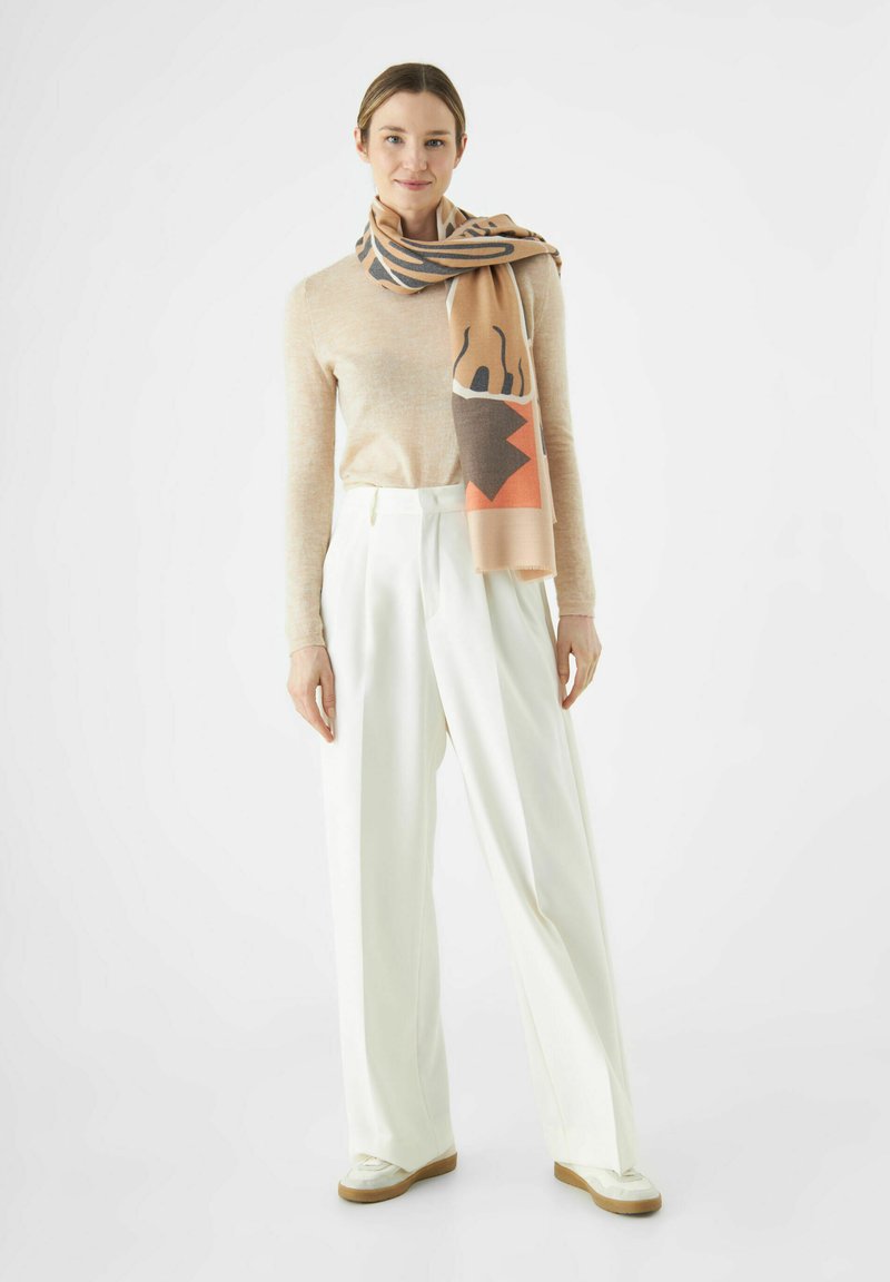 Pull en laine beige et pantalon large crème, assortis d'un foulard à motifs aux teintes naturelles et aux formes géométriques.