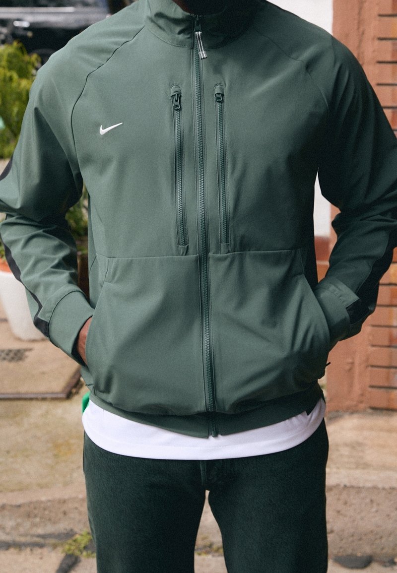 Vihreä vedenpitävä Nike-takki, jossa on korkea kaulus, vetoketjulliset rintataskut ja sivutaskut. Varustettu mustilla yksityiskohdilla ja sileällä pinnalla.
