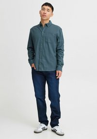 Mørk teal knapp-up skjorte med lange ermer og brystlomme, kombinert med mørk blå jeans og grå joggesko med svarte striper.