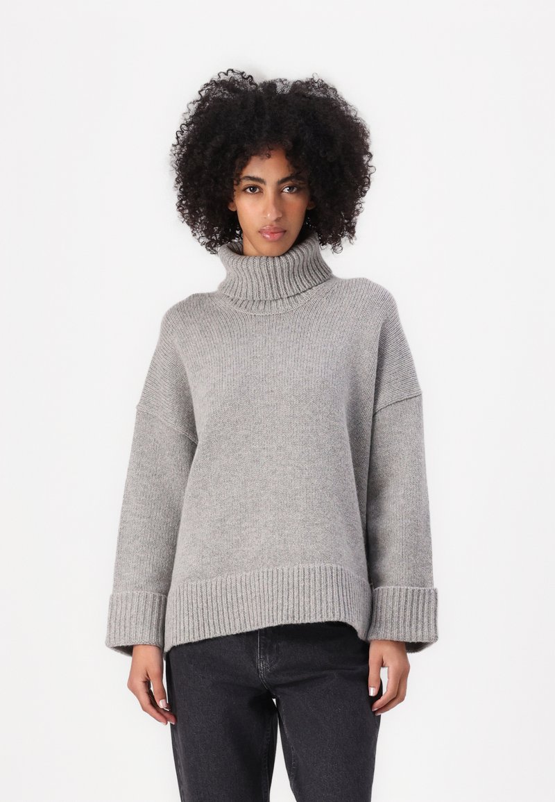 Suéter gris con un alto cuello de tortuga acanalado, diseño oversized y mangas anchas. Un acanalado grueso en el dobladillo y los puños añade textura.