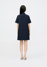 Mulher com cabelo preto longo, vestindo um vestido azul-marinho de manga curta e comprimento até ao joelho, e sapatos de salto bege, de pé de costas em um fundo branco.