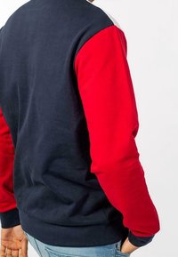 Marinblå och röd bomullströja med långa ärmar; baksidan har övre delen i hel marinblå och nedre ärmar i röd. Slät textur, avslappnad passform.