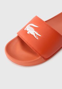 Orange slide sandal with textured footbed and white crocodile logo on the upper strap against a plain background.