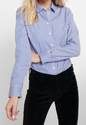 Kobieta ubrana w niebiesko-białą koszulę w paski z długimi rękawami i czarne spodnie, pozująca z jedną ręką zgiętą na talii.