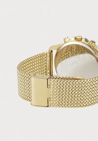 Le bracelet de montre en maille dorée présente un design tissé texturé et un fermoir poli. L'arrière est solide, réfléchissant la lumière avec une finition lisse.