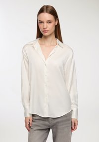 Blusa di seta bianca con bottoni sul davanti, colletto morbido a contrasto e maniche lunghe, abbinata a jeans in denim grigio chiaro.