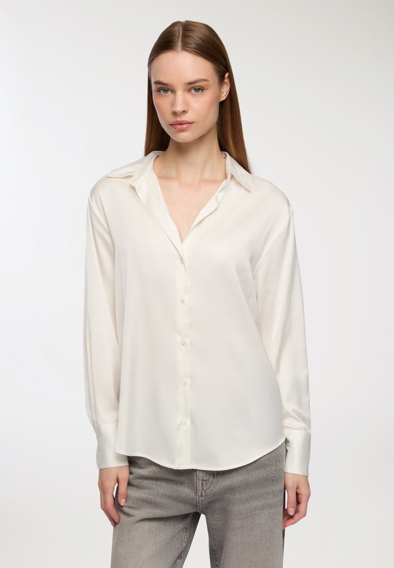 Blusa di seta bianca con bottoni sul davanti, colletto morbido a contrasto e maniche lunghe, abbinata a jeans in denim grigio chiaro.
