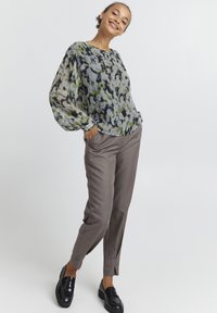 Blusa floreale trasparente in verde, grigio e blu navy, abbinata a pantaloni su misura color marrone chiaro e mocassini neri, con gambe affusolate.