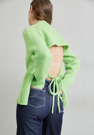 Persoană îmbrăcată cu un pulover tricotat deschis-verde, cu spatele gol, legat în talie, asortat cu blugi de culoare albastră închis, cu cusături albe pe buzunare.