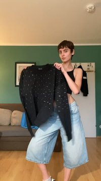 Suéter preto com detalhes em prata espalhados, com um corte solto e ombros caídos, segurado por uma pessoa vestindo um top cropped preto.
