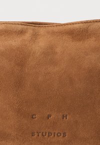 Borsa in suede marrone con una texture morbida. Presenta il logo impresso "C P H STUDIOS" sul fronte. Design semplice e rettangolare senza accessori.