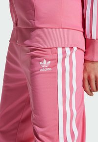 Ružový teplákový komplet s textúrovaným povrchom. Má biele trojprúžkové akcenty po stranách a vyšívané logo Adidas na nohe nohavíc.