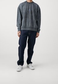 Sudadera gris con patrón repetitivo en negro del logo "HUGO", cuello redondo y puños acanalados, combinada con jeans azul oscuro y zapatillas blancas.
