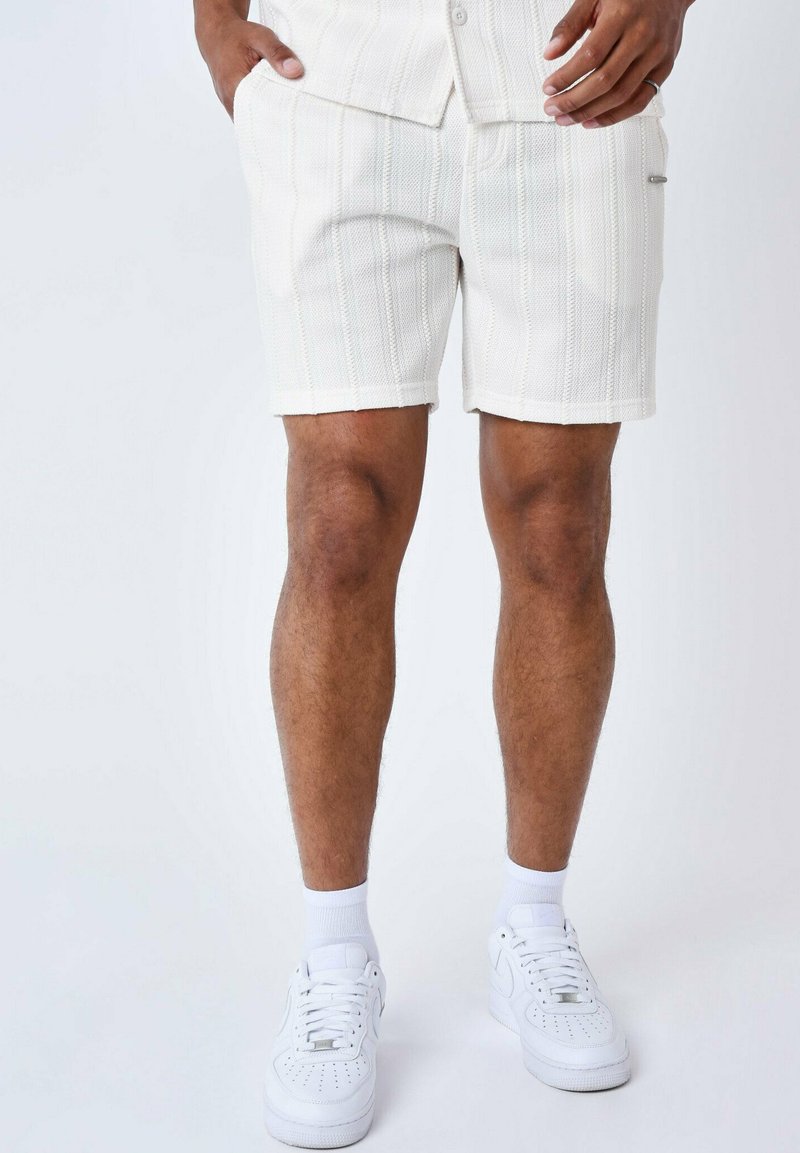 Bermuda bianche con texture e righe verticali, dotate di tasche laterali e una leggera lucentezza. Abbinati a sneakers bianche e calzini alla caviglia.