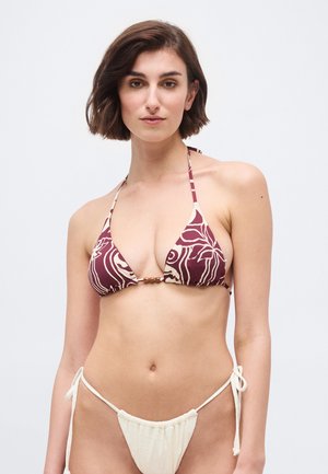 Νεαρή γυναίκα με κοντά καστανά μαλλιά, φοράει ένα μαγιό πάνω μέρος με μαροκινό μοτίβο και ένα μονόχρωμο κρεμ κάτω μέρος με κορδόνια στο πλάι, στέκεται μπροστά σε απλό φόντο.