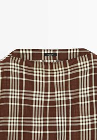 Tissu à carreaux brun et beige avec un col rectangulaire et une petite étiquette noire portant l’inscription "Massimo Dutti Made in China" à l’encolure.