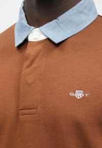 Bruin poloshirt met een chambray kraag, knoopsluiting en geborduurd logo. Heeft een geribbelde structuur en een contrasterend kraagmateriaal.