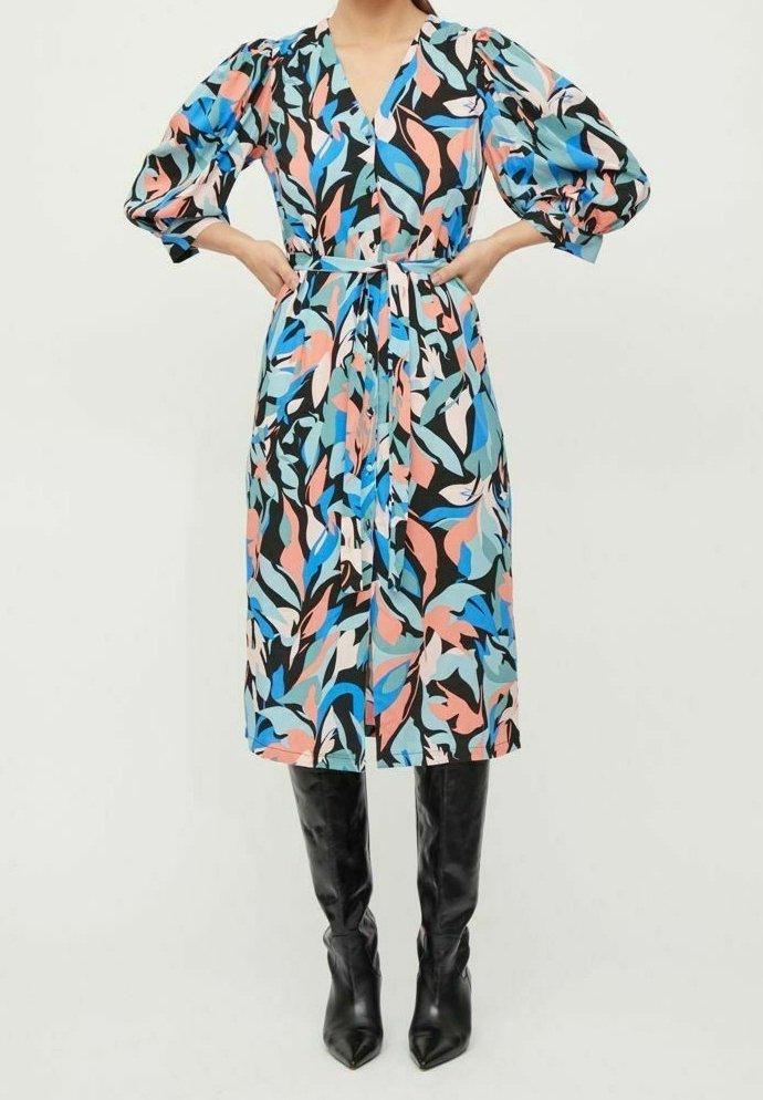 Femme portant une robe ceinturée longueur genou avec des motifs abstraits de feuilles bleues, roses, noires et blanches, associée à des bottes noires pointues montant jusqu’aux genoux.
