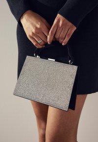 Silberne, verzierte Handtasche mit strukturiertem Design und schwarzen Akzenten, ausgestattet mit einem Tragegriff und einer strukturierten Oberfläche.