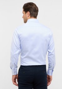 Camicia a maniche lunghe azzurro chiaro con una texture liscia, colletto a punta e polsini con bottoni, vista da dietro, abbinata a pantaloni scuri.