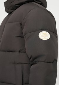 Sorte puffjakke med tekstureret stof, quiltet design, hætte og en cremet logo-patch på venstre ærme.