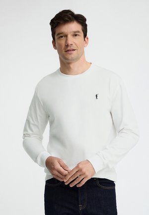 Mann trägt ein weißes Langarmhemd mit einem kleinen schwarzen Golfer-Logo auf der Brust, kombiniert mit dunkelblauen Jeans.