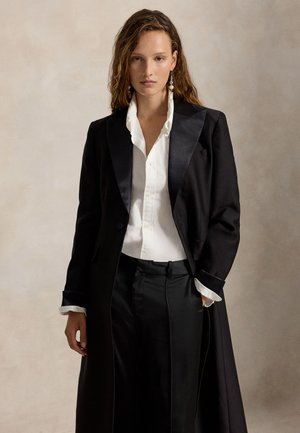 Femme aux cheveux ondulés porte un long manteau noir, une chemise blanche boutonnée, un pantalon noir et des boucles d'oreilles pendantes, debout devant un fond neutre.