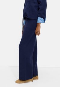 Marineblaue weite Hosen aus weichem Stoff, kombiniert mit einem marineblauen Oberteil mit hellblauen Ärmeln und einer Vordertasche. Braune Schuhe runden das Outfit ab.