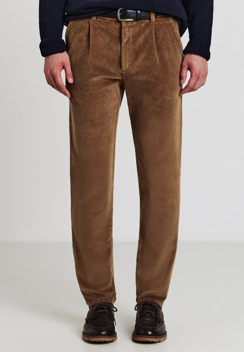 Mann trägt braune Cordhosen mit vorderen Falten, schwarzen Gürtel, marineblauen Pullover und dunkelbraunen Lederschuhen und steht auf einem hellen Boden.