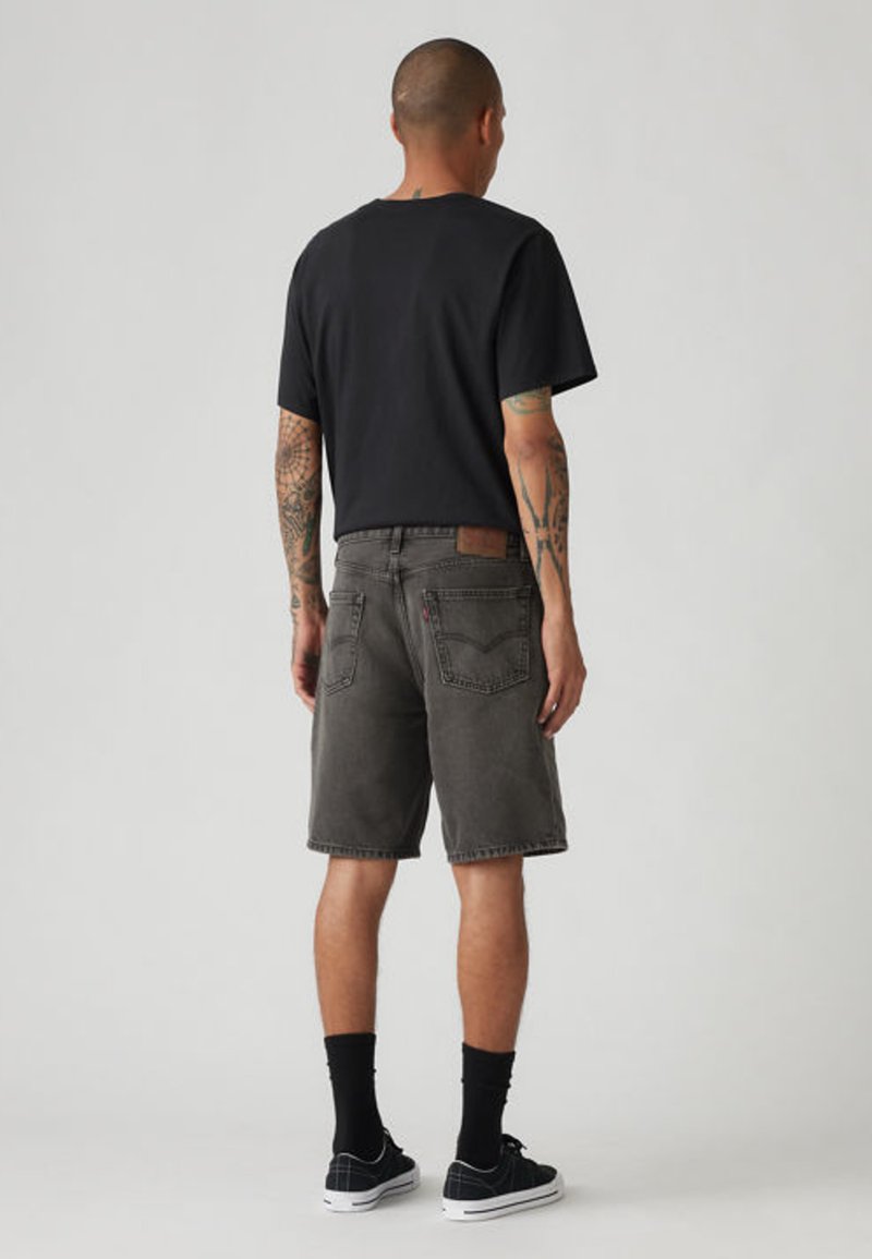 Mand med tatoveringer iført sort t-shirt, falmede sorte denimshorts, sorte sokker og sorte sneakers, stående med ryggen til mod en ensfarvet baggrund.