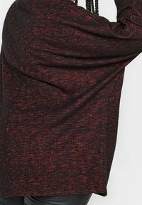 T-shirt à manches longues en tissu texturé noir et rouge avec une coupe décontractée et des côtés drapés, présentant un ourlet arrondi et des manches larges.