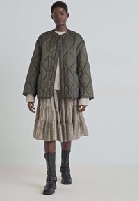Olive green quilted jacket with a round neck, layered over a beige knit sweater and plaid tiered skirt, paired with black knee-high boots.