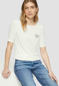 Witte T-shirt met korte mouwen van zachte stof met een ronde hals en een strass-strikontwerp op de linkerkant van de borst, gecombineerd met blauwe jeans.